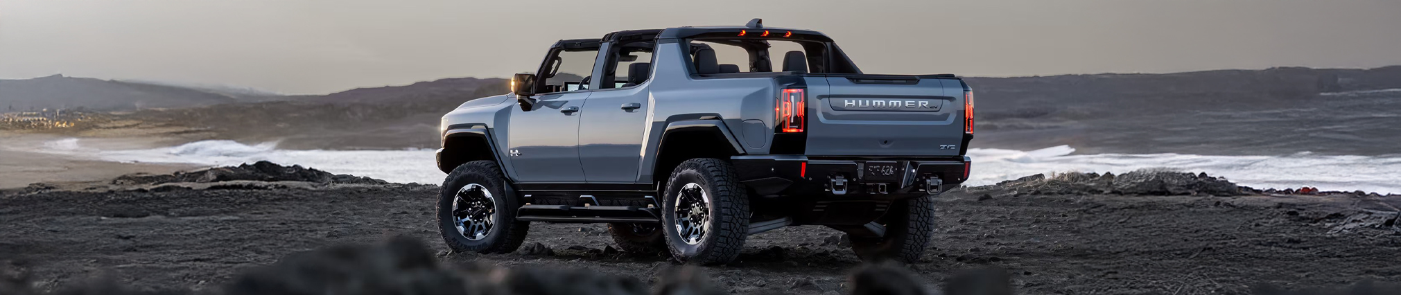 Back-side view of the 2025 GMC Hummer EV Pickup Truck parked near a body of water by a rocky terrain.