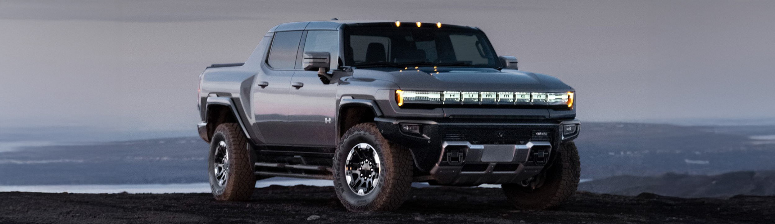 Front-side view of the 2025 GMC Hummer EV Pickup Truck parked near a body of water by a rocky terrain.