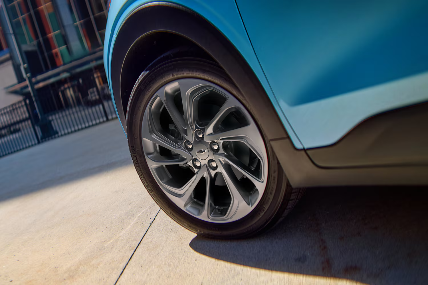 A Close-Up of the Front Wheel of a Light Blue 2027 Chevrolet Bolt, Showing the Multi-Spoke Aluminum Wheel with a Chrome Chevrolet Bowtie in the Center.