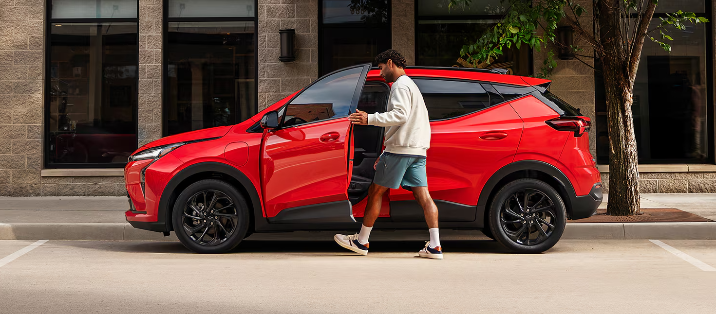 The Side View of a Red 2027 Chevrolet Bolt, with a Man Getting into the Driver's Side Door on a City Street.